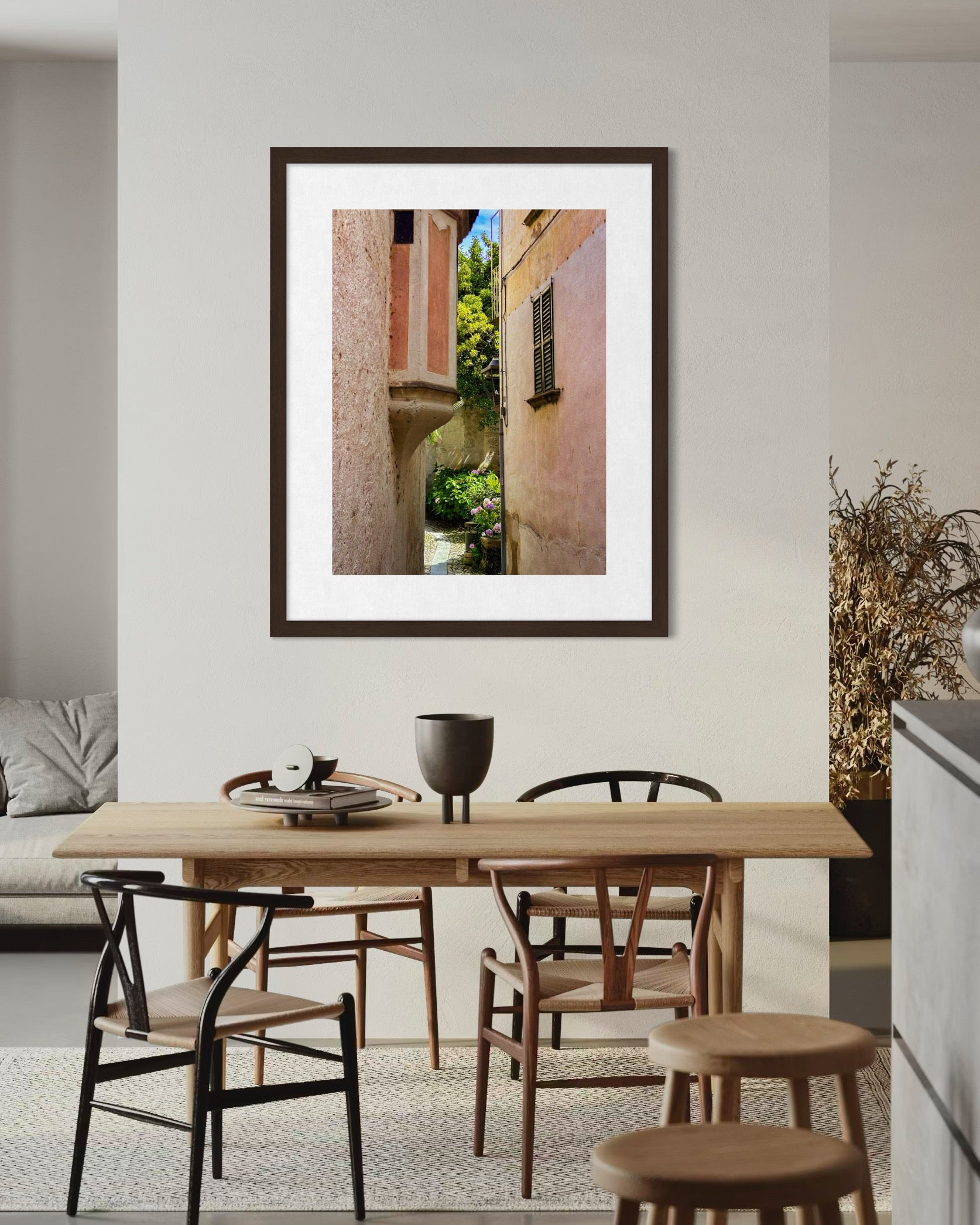 “Hidden Passage on Lago di Maggiore” fine art print by Leslie Brashear Photography, depicting a narrow alley with stucco buildings and greenery, hangs above a light wood dining table in a modern dining area.