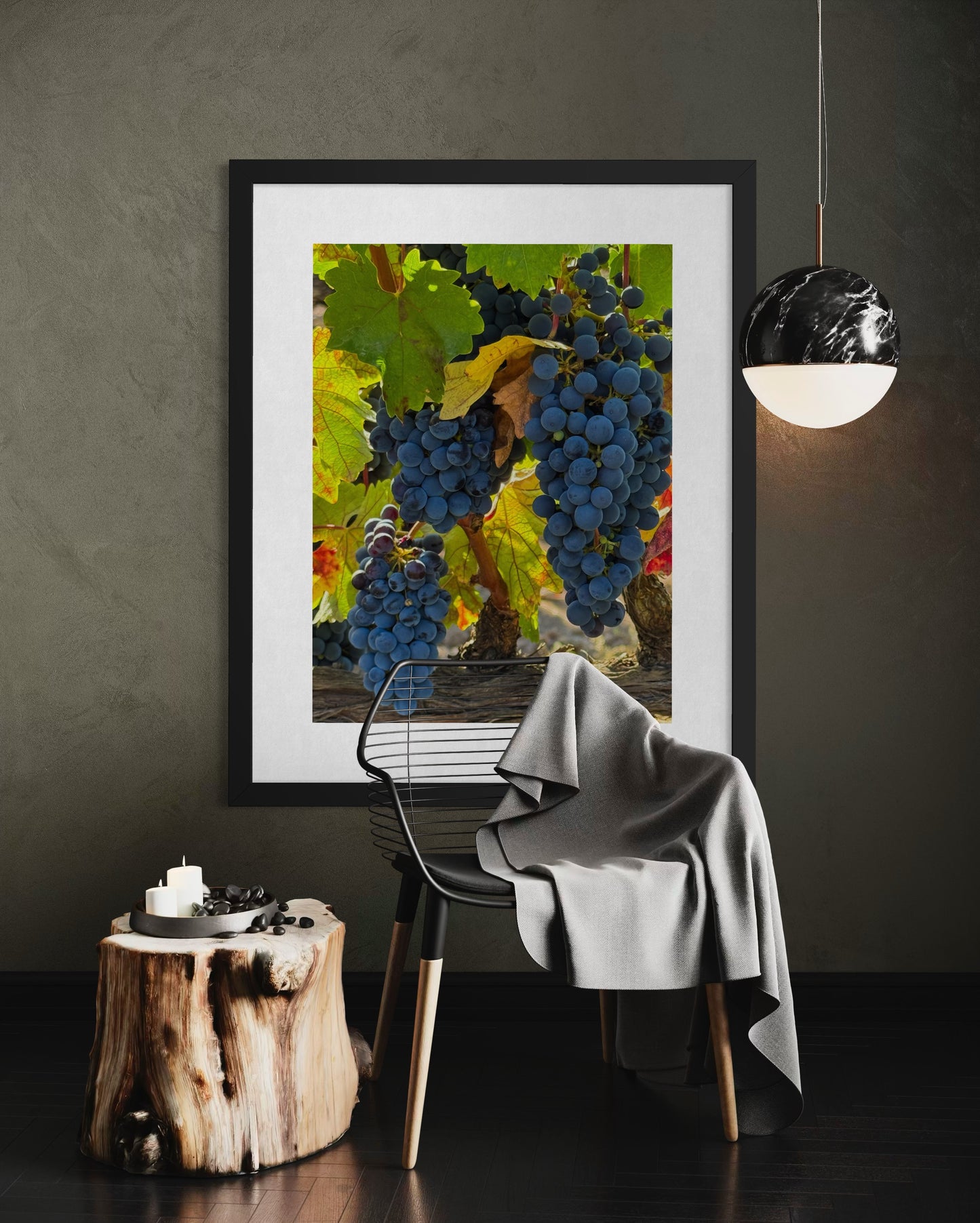 A Trio of Cabernet Sauvignon Grape Clusters - Fine Art Print by Leslie Brashear Photography hangs above a modern chair with a gray blanket, beside a wooden stump table and a glowing pendant lamp on a dark wall.