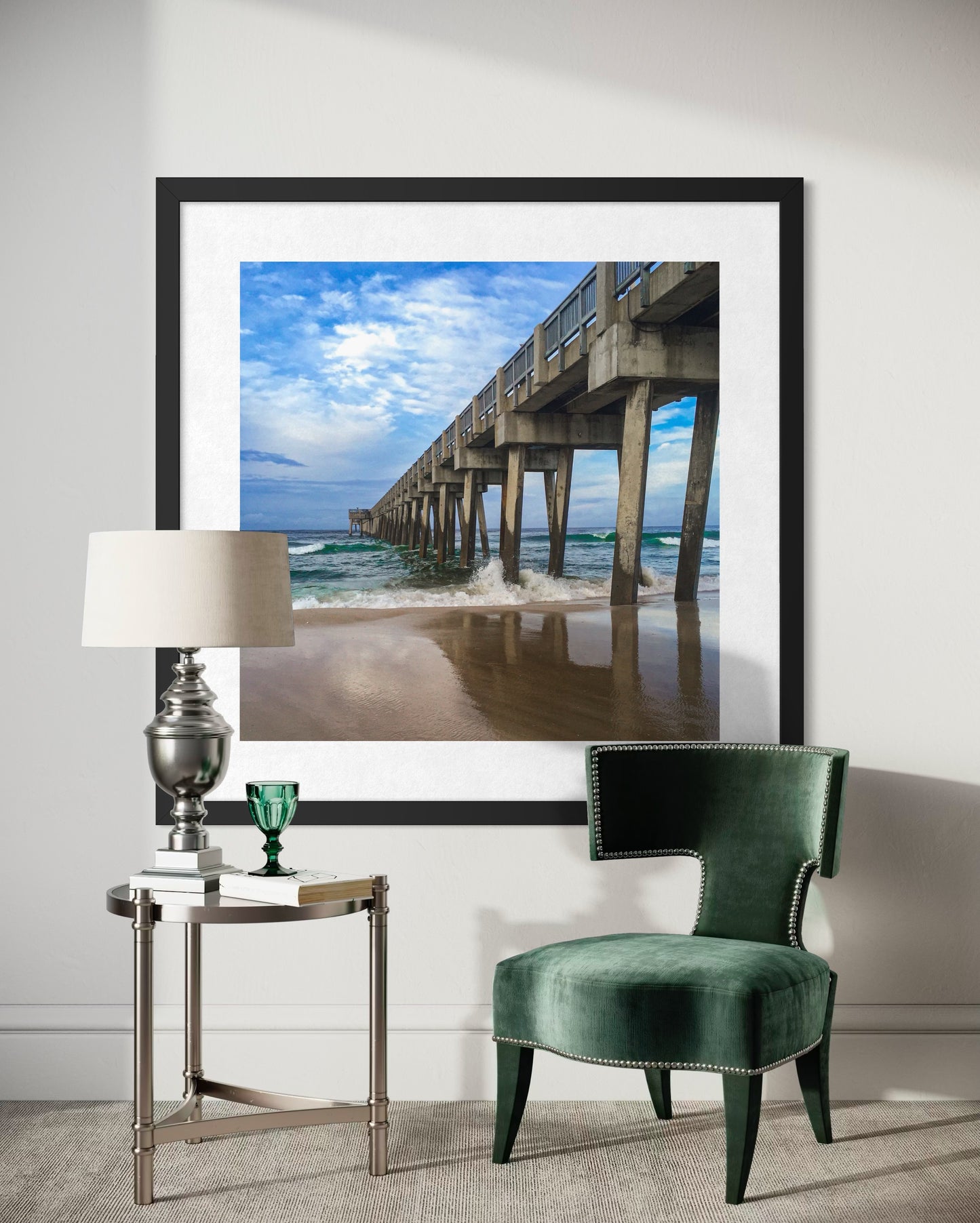 A framed "Pier Reflection - Fine Art Print" by Leslie Brashear Photography hangs on a white wall above a small table with a lamp, glass, and books, next to a green velvet chair.