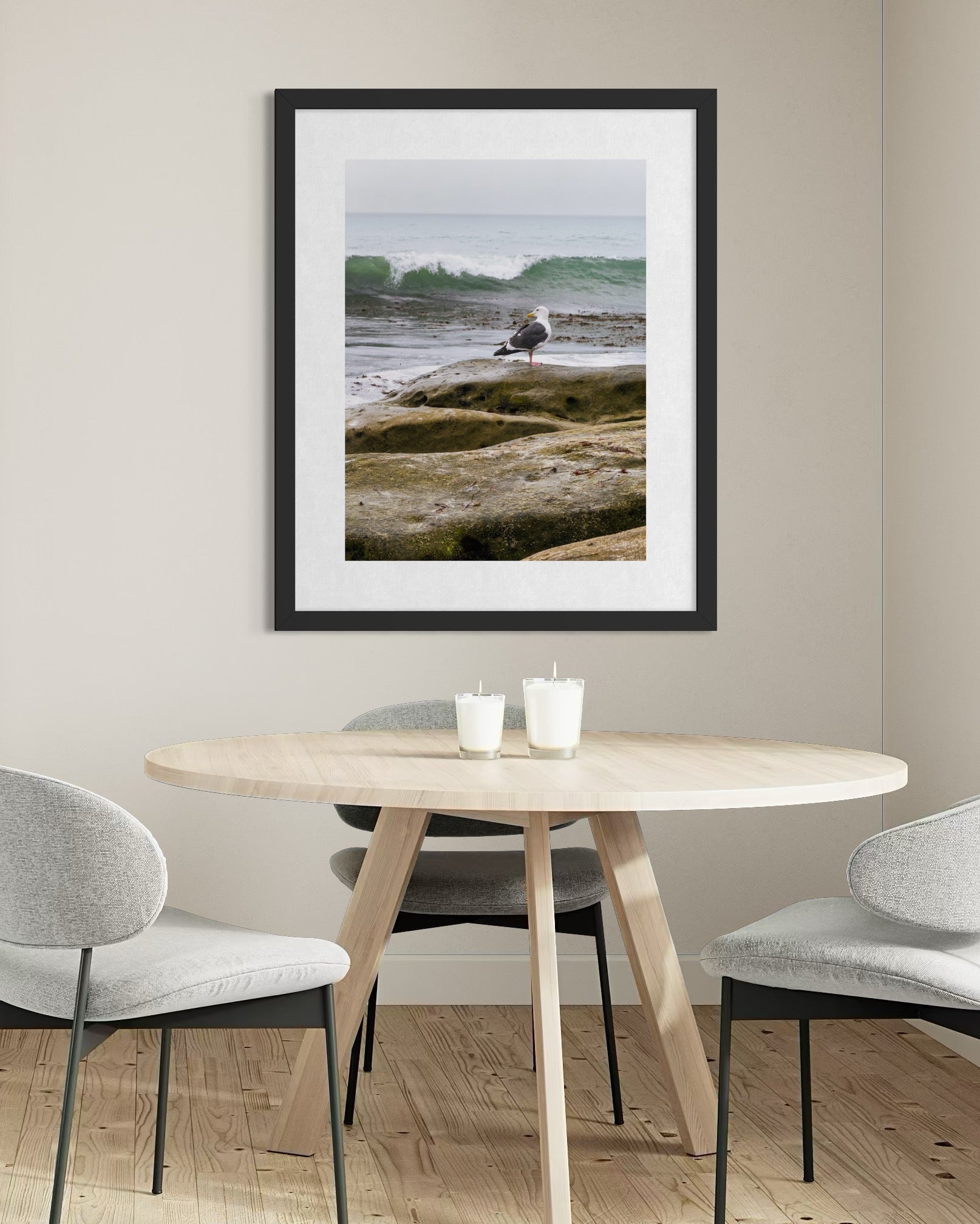 A modern dining room features a round wooden table, three gray chairs, two candles, and "Seagull on the Rocky Shoreline - Fine Art Print" by Leslie Brashear Photography—adding an elegant touch of coastal artistry to the wall.