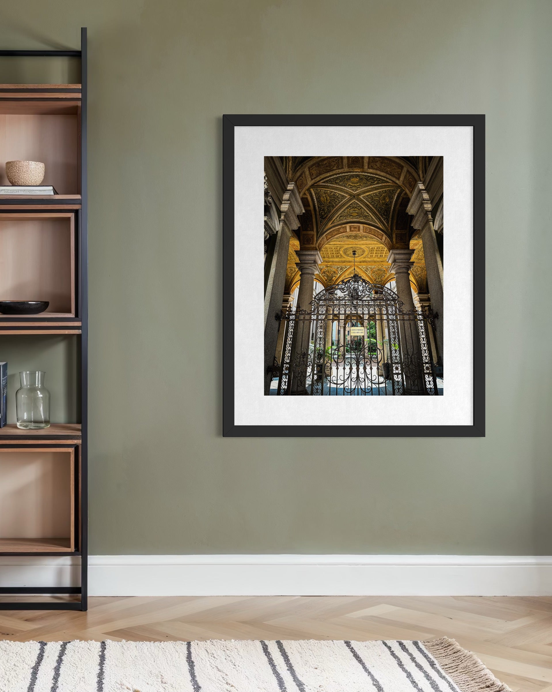 The Grand Entrance in Milan - Fine Art Print by Leslie Brashear Photography, featuring an ornate arched doorway with iron gates, is displayed on a green wall next to wooden shelves in a modern room with a light rug and hardwood floor.