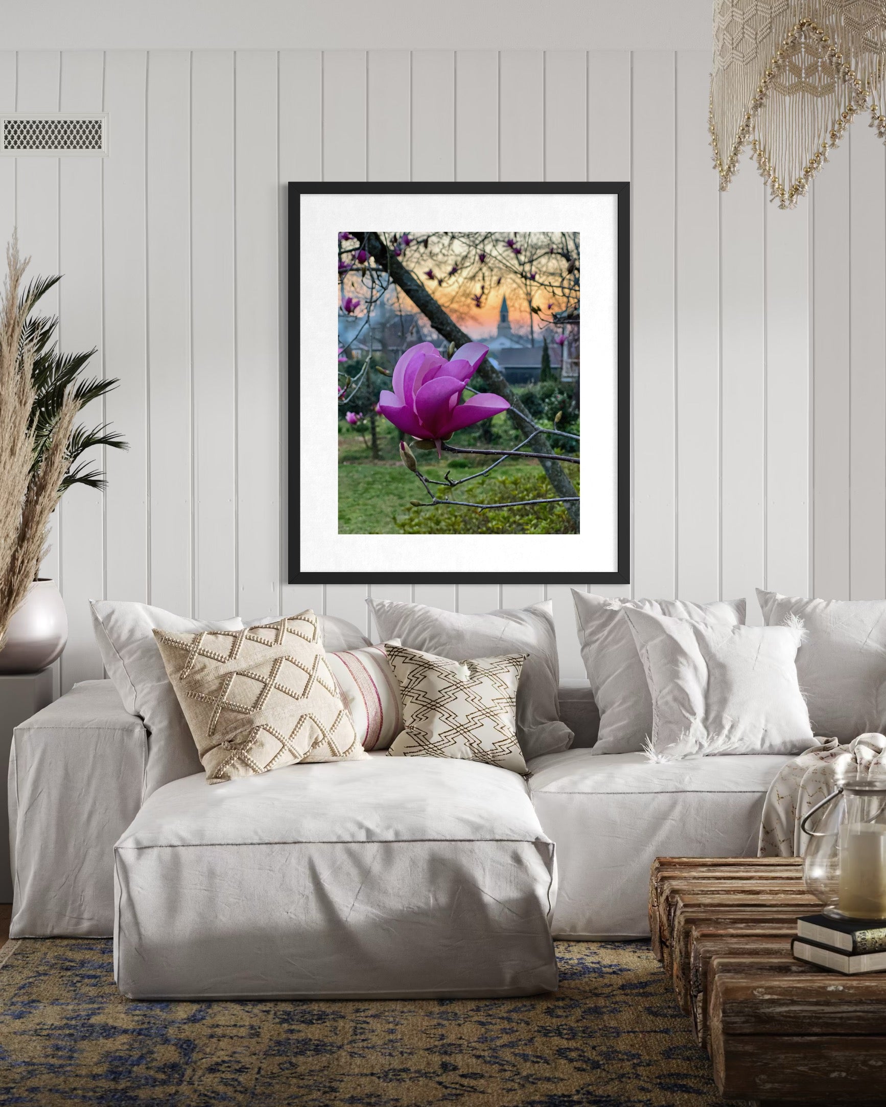 A living room with a white sofa, patterned cushions, a wooden coffee table, and the "Pink Magnolia - Fine Art Print" by Leslie Brashear Photography displayed on a white paneled wall.