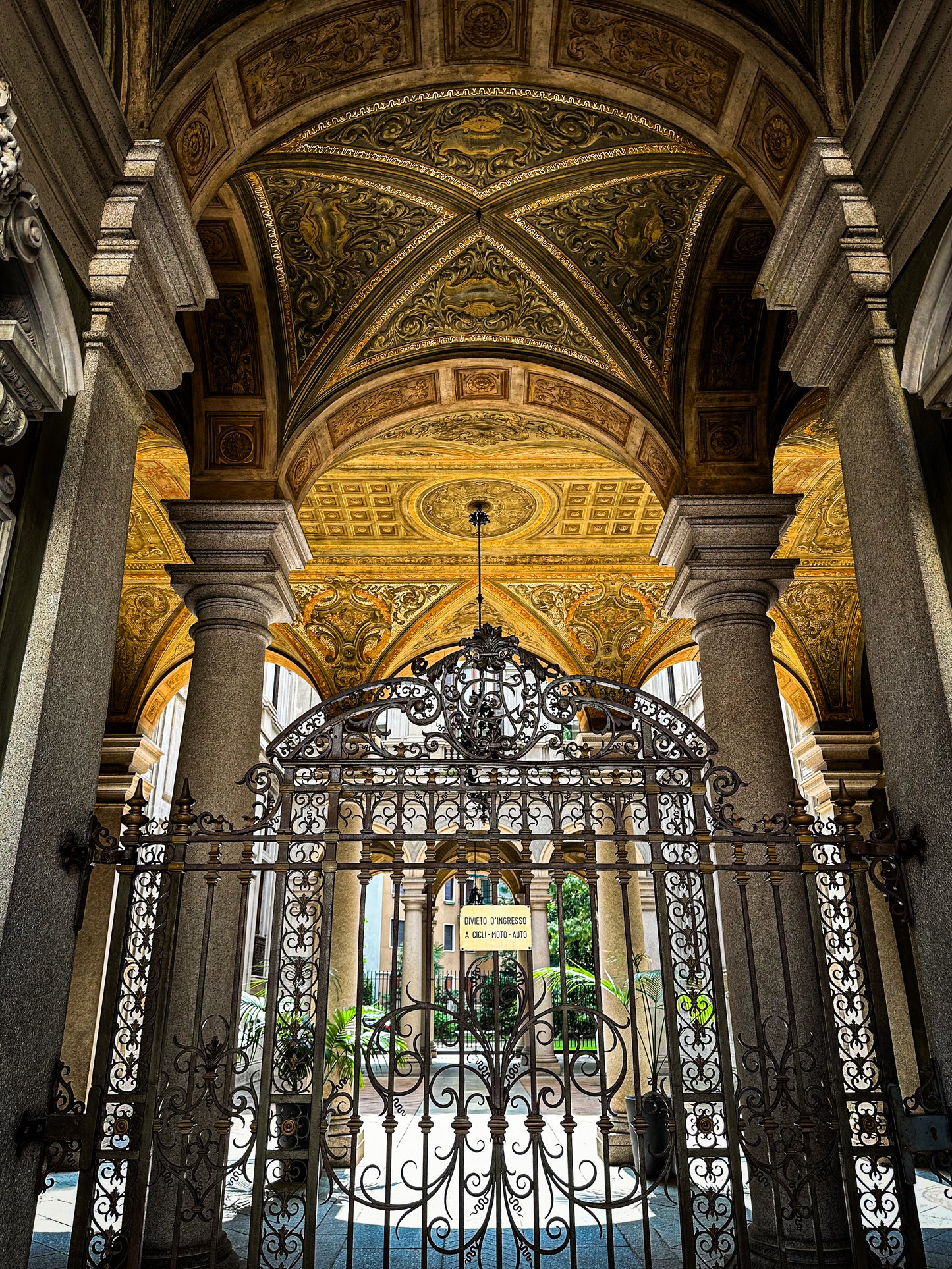 Discover "Grand Entrance in Milan - Fine Art Print" by Leslie Brashear Photography, featuring an ornate arched ceiling with gold designs, stone columns, and an intricate iron gate opening to a courtyard—ideal for architecture lovers.