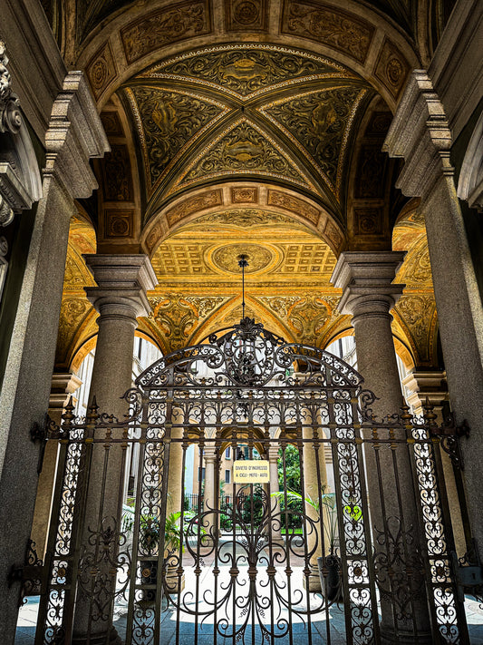 Discover "Grand Entrance in Milan - Fine Art Print" by Leslie Brashear Photography, featuring an ornate arched ceiling with gold designs, stone columns, and an intricate iron gate opening to a courtyard—ideal for architecture lovers.