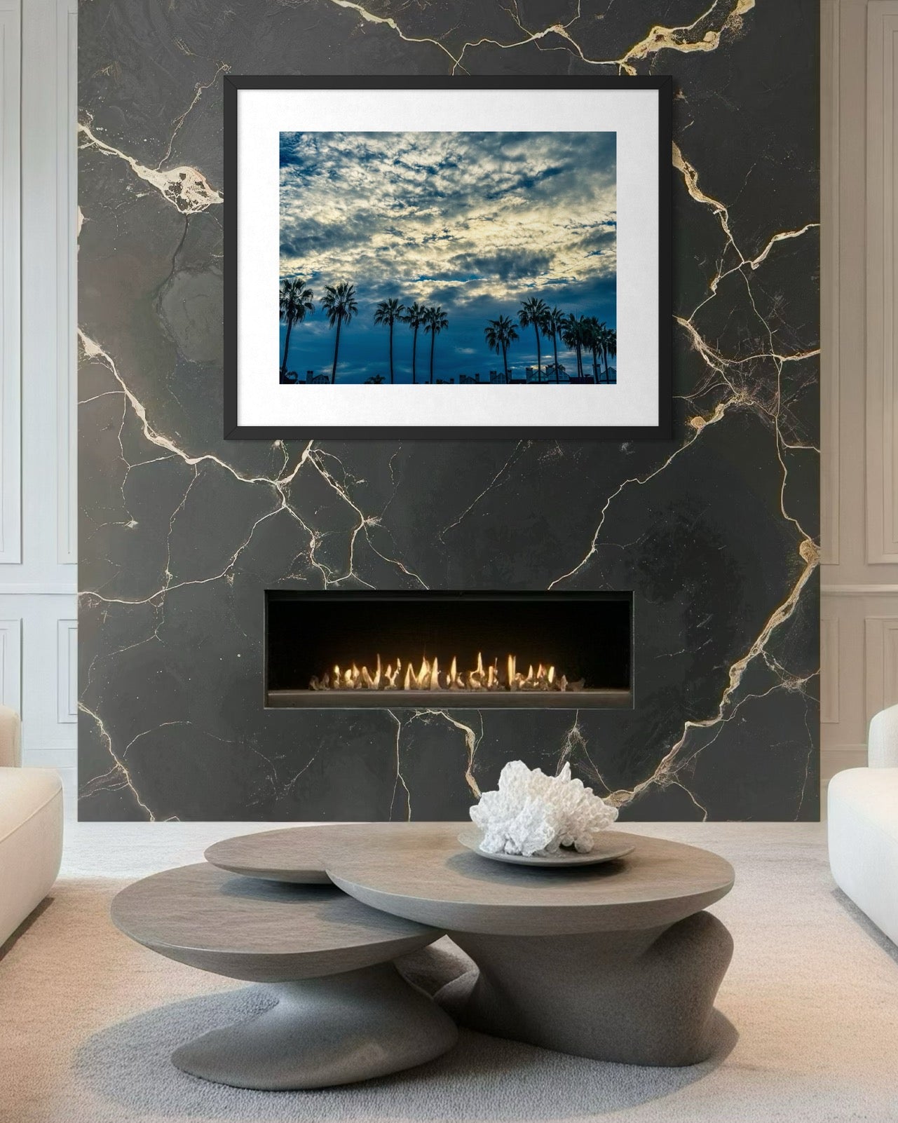 A modern living room features a round coffee table, marble wall, built-in fireplace, and "Silhouettes Under a Dramatic Sky" fine art print by Leslie Brashear Photography above the mantel, inspired by a San Diego sunset.