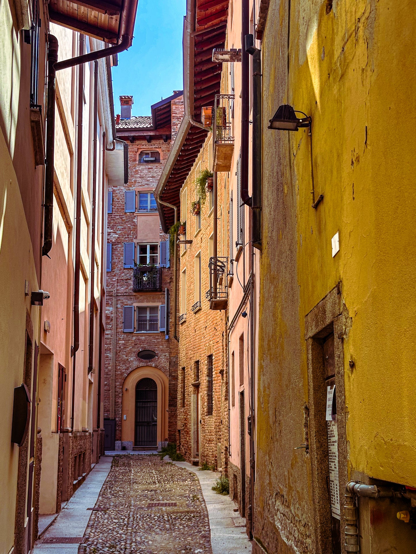 Alleyway in Pavia - Fine Art Print by Leslie Brashear Photography captures a charming cobblestone alley with yellow and brick buildings, balconies, and an arched doorway under a blue sky—ideal Italy wall art.