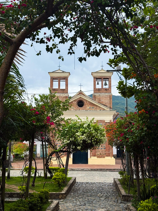 Charming Church - Fine Art Print by Leslie Brashear Photography features a brick church with twin bell towers framed by flowering plants and greenery along a cobblestone path, capturing classic Colombian architecture.