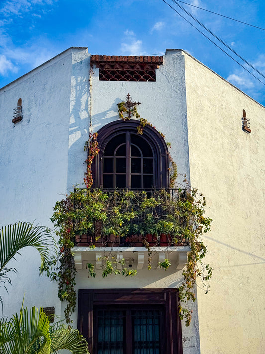 The "Charming Corner Balcony - Fine Art Print" by Leslie Brashear Photography captures a white building with an arched window and wrought iron balcony adorned with plants, embodying the charm of Guadalajara architecture.
