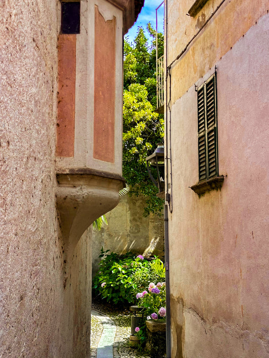 Hidden Passage on Lago di Maggiore - Fine Art Print by Leslie Brashear Photography captures a sunlit alley with a garden and pink hydrangeas, evoking the charm of Italian lakeside towns—ideal for Lombardy travel photography lovers.