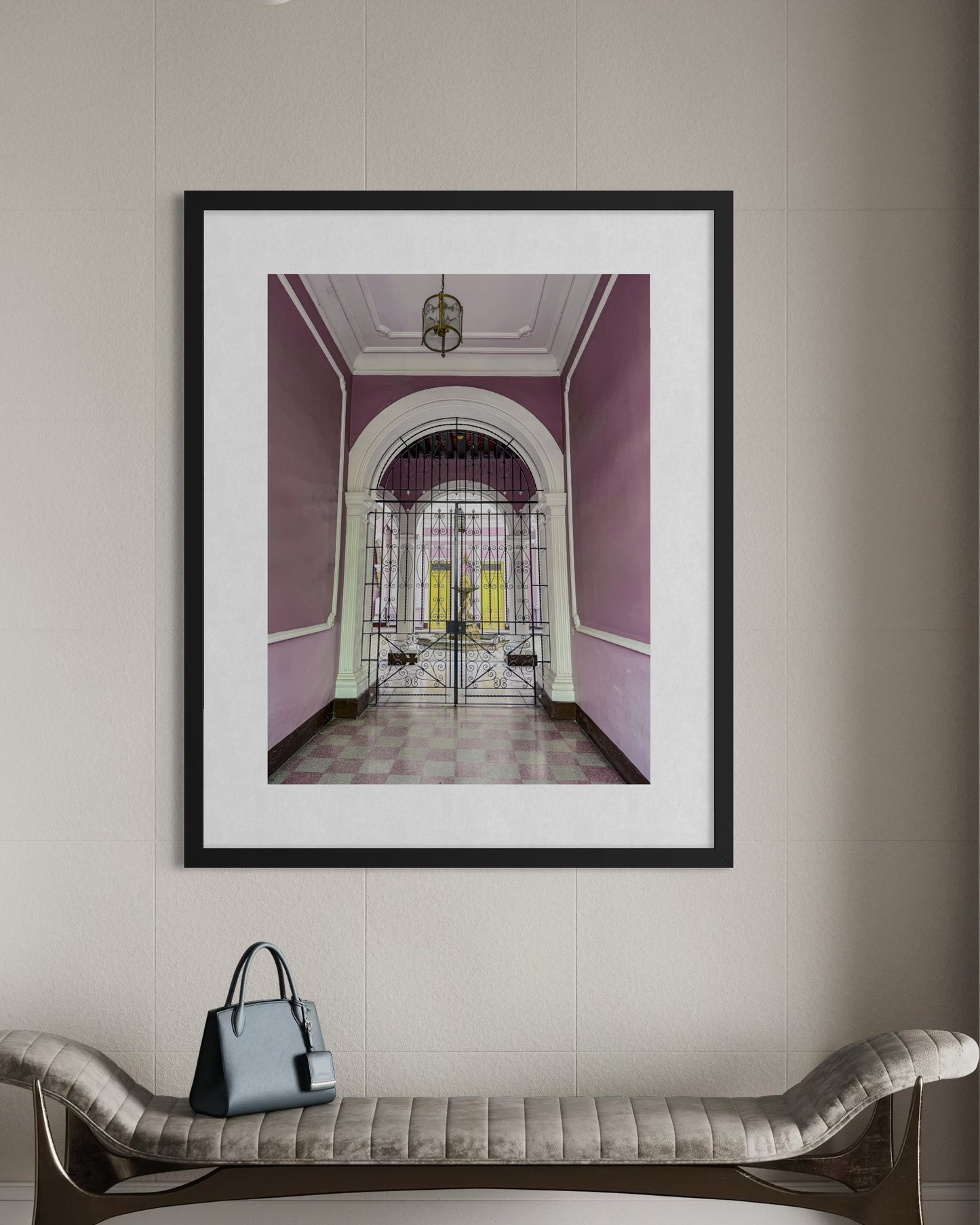 A Courtyard Through an Ornate Gate fine art print by Leslie Brashear Photography hangs on a beige wall above a cushioned bench topped with a gray handbag.