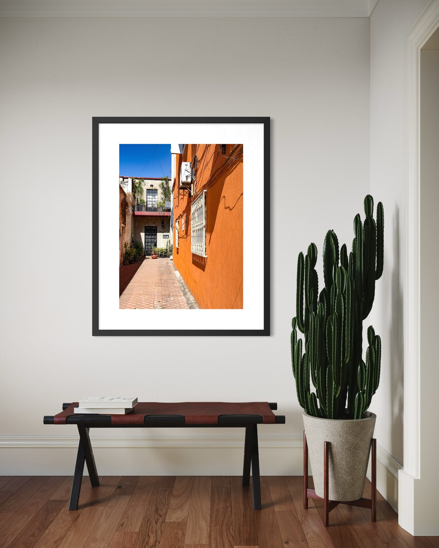 The "Sunlit Urban Alleyway - Fine Art Print" by Leslie Brashear Photography features an orange building and brick walkway, displayed above a bench with a tall potted cactus beside it on a wooden floor.