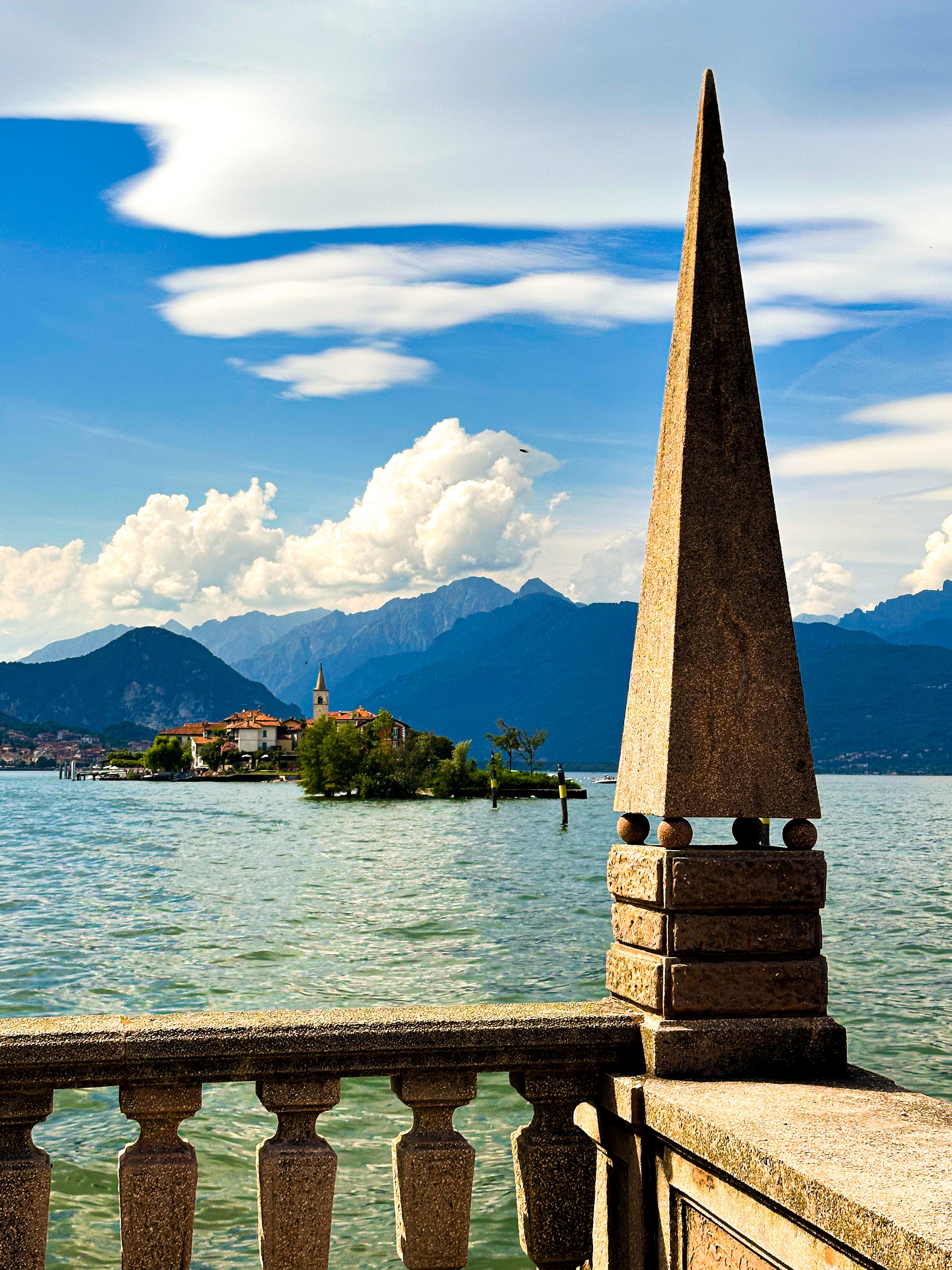 The Lago di Maggiore - Fine Art Print by Leslie Brashear Photography features a stone railing and pointed pillar overlooking a scenic lake, picturesque island, and distant mountains beneath a partly cloudy sky.