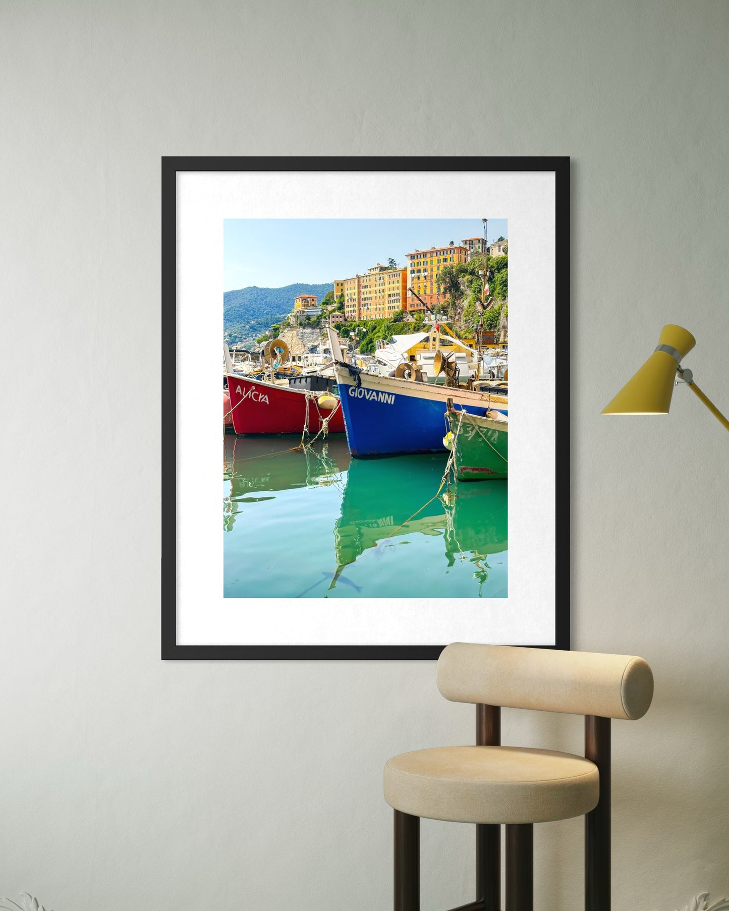 Reflections - Fine Art Print by Leslie Brashear Photography, featuring colorful boats in Camogli harbor, displayed on a white wall above a beige chair and next to a yellow lamp.