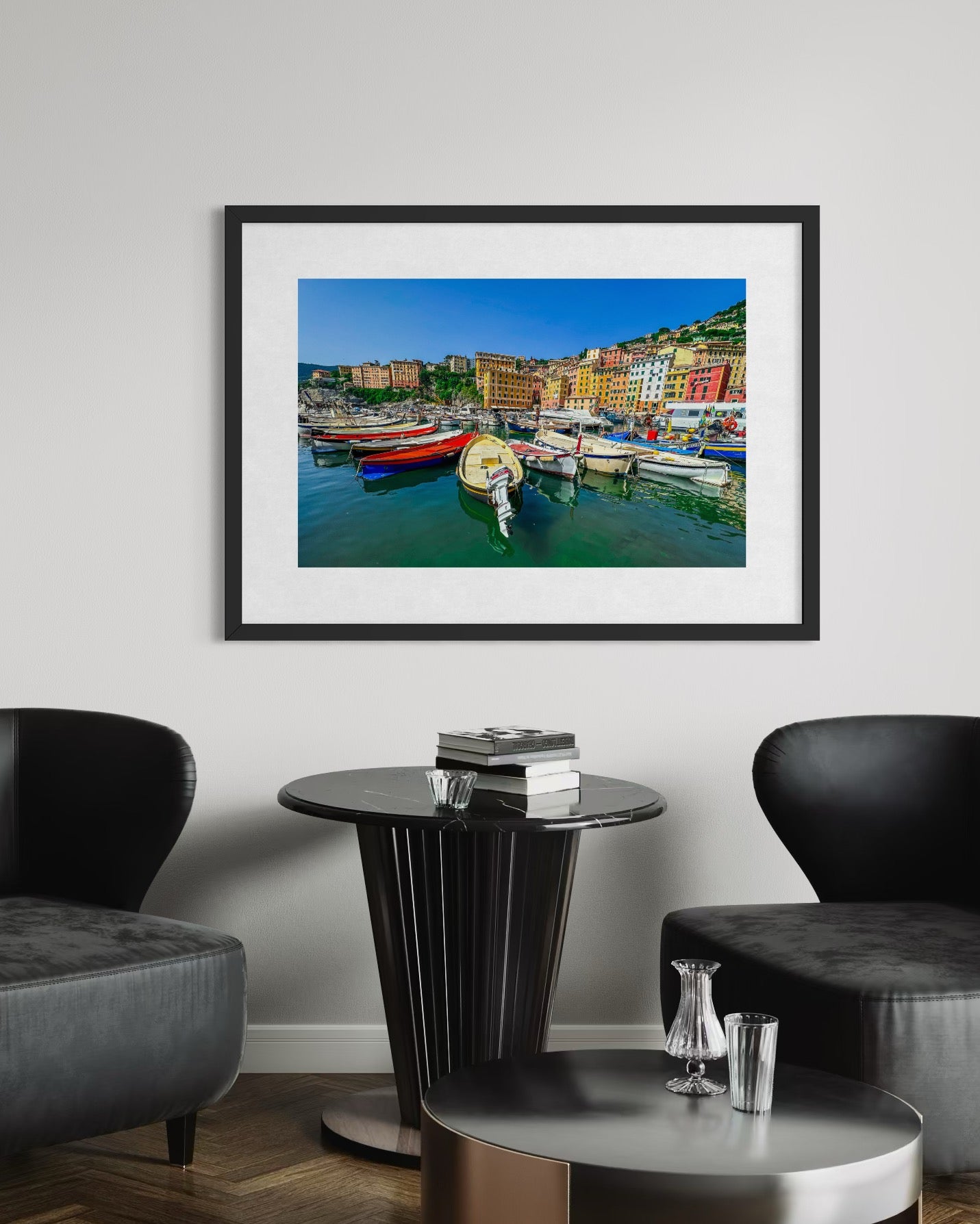 A Camogli Harbor Fine Art Print by Leslie Brashear Photography hangs on a white wall above a round black table between two black chairs, showcasing colorful boats along Italy’s Ligurian coast.