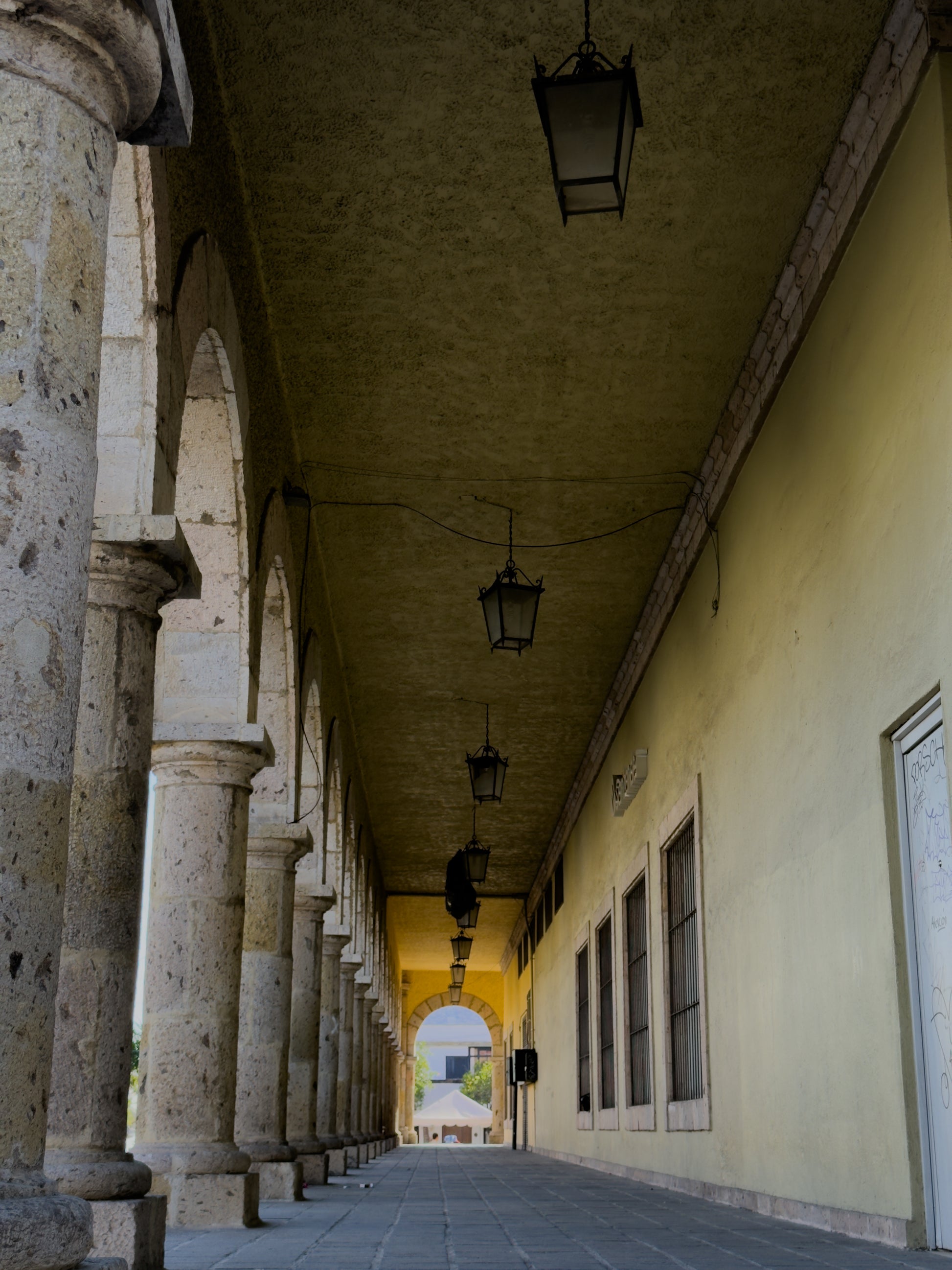 The Zapopan Stone Colonnade Passageway Fine Art Print by Leslie Brashear Photography showcases a classic stone archway corridor with lanterns, columns on the left, and pale yellow walls leading to an open arch.