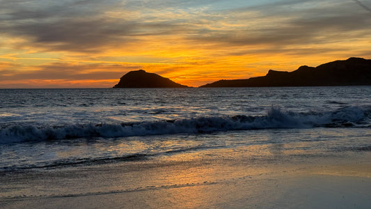 Sunset Over the Coast of Mazatlan by Leslie Brashear Photography captures a vibrant dusky sky, waves breaking along the shore, and dark silhouettes of coastal cliffs on the horizon.