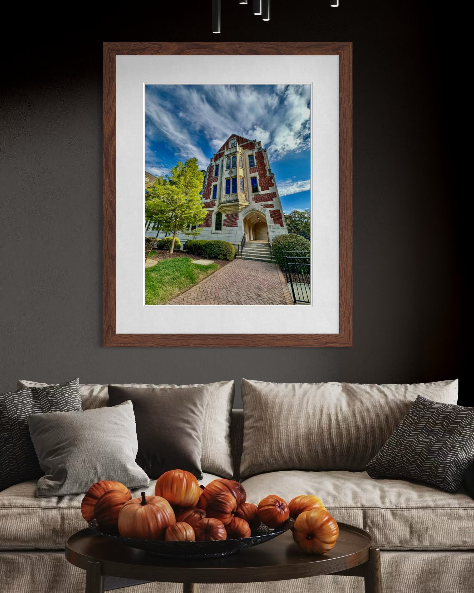 A framed "Agnes Scott College" photo by Leslie Brashear Photography, featuring a red brick Gothic Revival building, hangs on a dark wall above a beige sofa; decorative pumpkins fill a bowl on the coffee table.