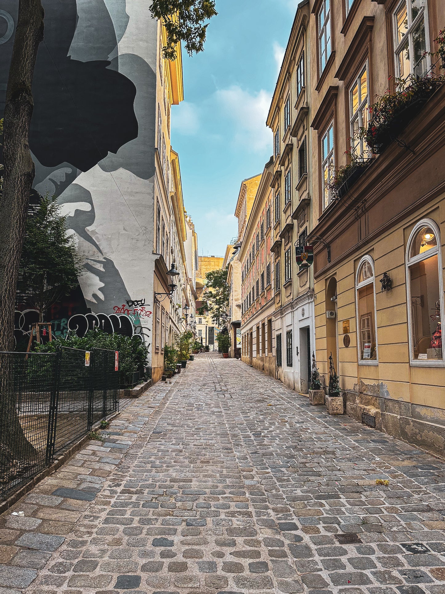 Quiet Alley in Vienna - Fine Art Print by Leslie Brashear Photography captures a cobblestone street framed by historic Vienna architecture and a vibrant mural, all beneath a clear blue sky.