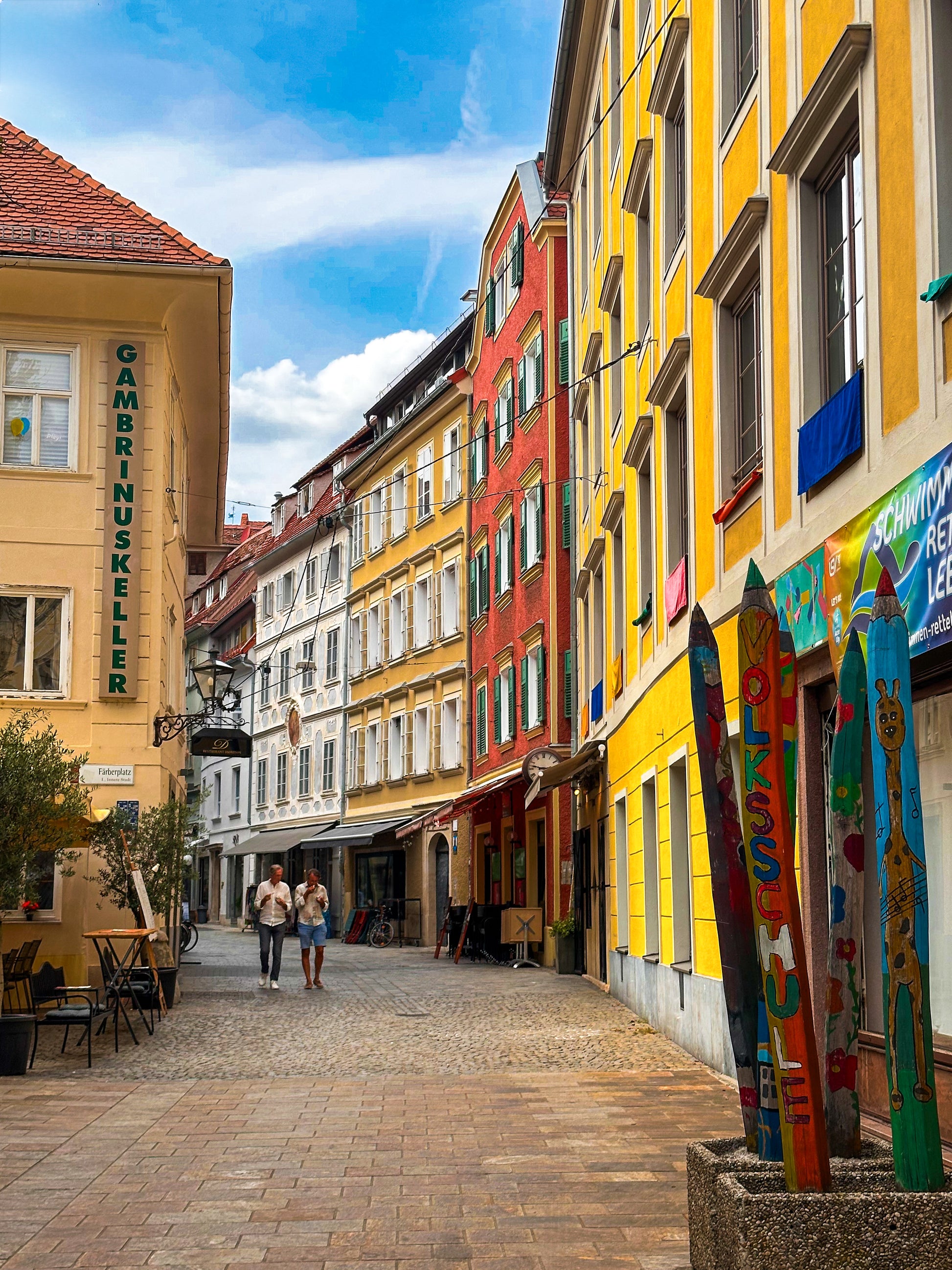 “Walking in Graz - Fine Art Print” by Leslie Brashear Photography captures two people strolling past upright painted wooden skis and colorful buildings on a cobblestone street under a partly cloudy sky in Austria.