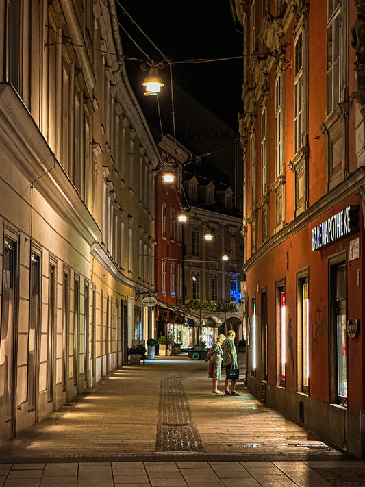 Window Shopping - Fine Art Print by Leslie Brashear Photography captures a narrow, well-lit cobblestone Graz alley at night, with people near a pharmacy and classic European architecture lining the residential buildings.