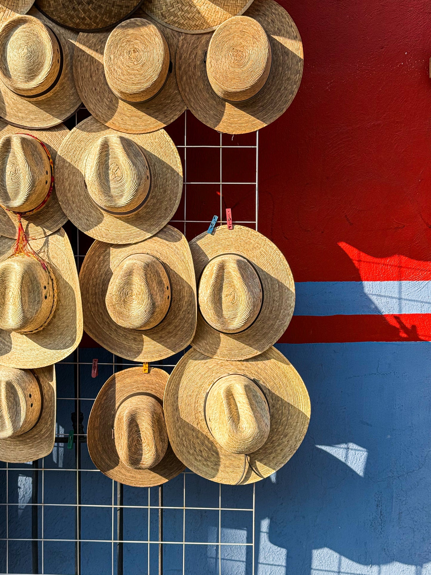 “Straw Hats - Fine Art Print” by Leslie Brashear Photography features Mexican straw hats hanging on a metal grid against a vivid red and blue wall—sunlit shadows highlight this striking piece of cultural decor.