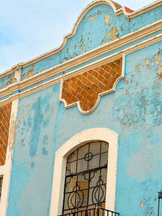 Blue Facade in Puebla - Fine Art Print by Leslie Brashear Photography captures a weathered blue building with peeling paint, an arched window with ironwork, and brown geometric accents—an embodiment of Mexican facade art.