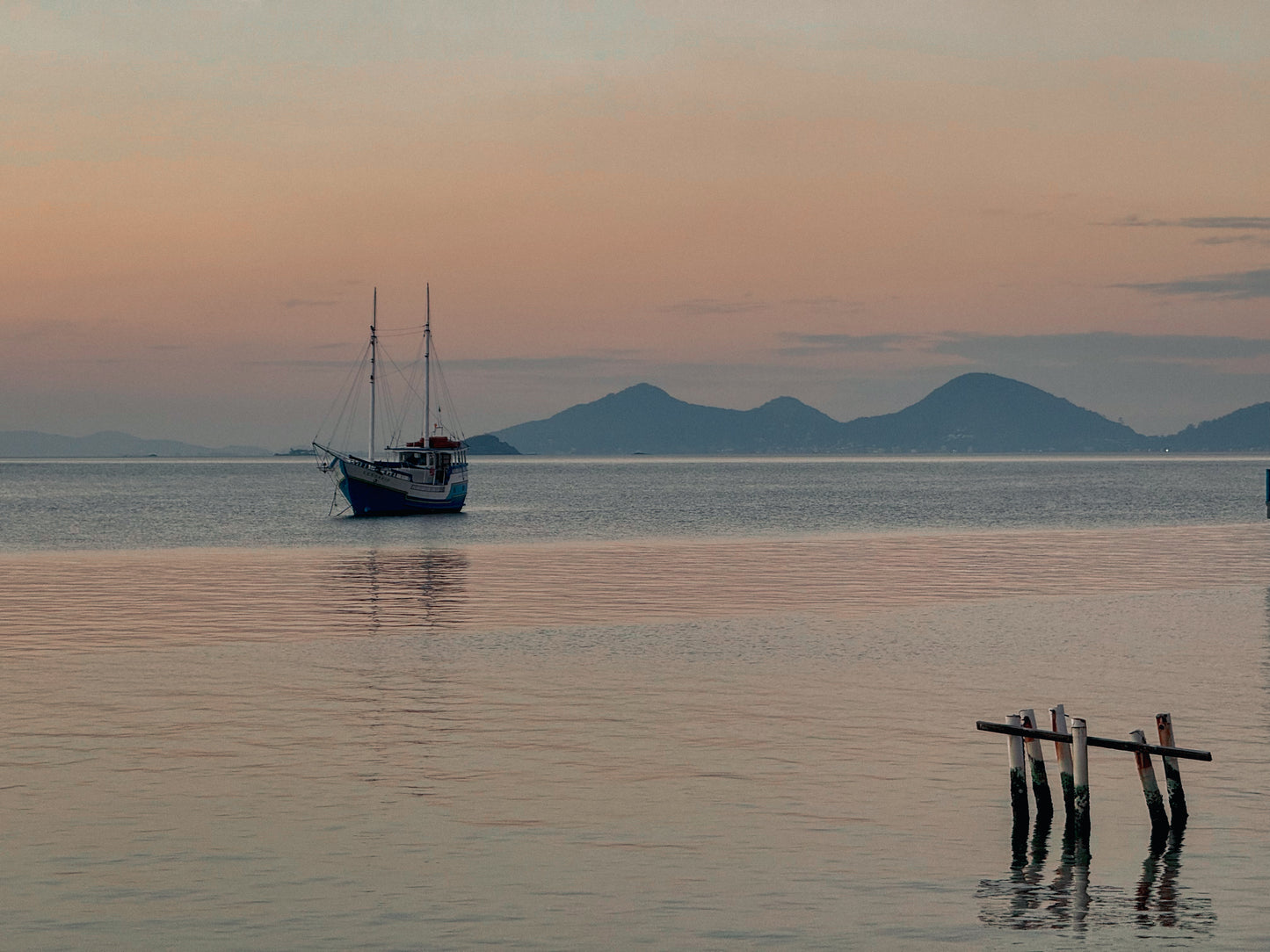 Anchored in Tranquility - Fine Art Print by Leslie Brashear Photography captures a serene sunset illuminating a sailboat on calm Florianópolis waters, with distant hills and weathered wooden posts adding to the peaceful atmosphere.