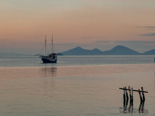 Anchored in Tranquility - Fine Art Print by Leslie Brashear Photography captures a serene sunset illuminating a sailboat on calm Florianópolis waters, with distant hills and weathered wooden posts adding to the peaceful atmosphere.
