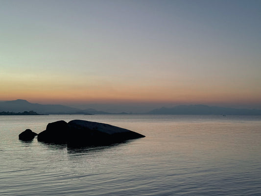 Capture tranquil coastline beauty with "Twilight Serenity Over Still Waters"—a fine art print by Leslie Brashear Photography, featuring calm water, large foreground rocks, gentle ripples, and distant mountains at dusk.