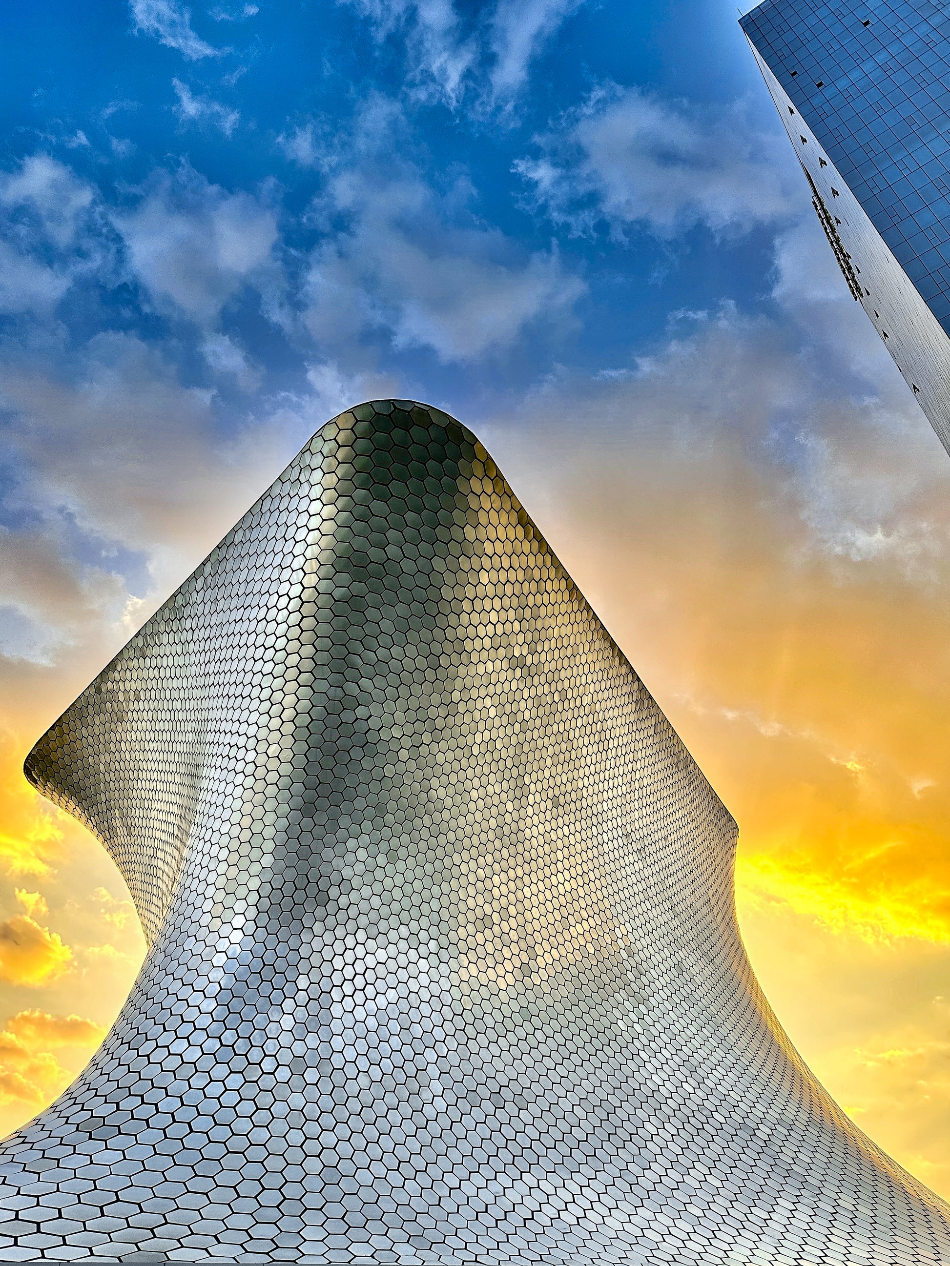 Soumaya Museum at Sunset - Fine Art Print by Leslie Brashear Photography captures the curved, honeycomb-patterned Soumaya Museum in Mexico City at sunset, with a tall glass building nearby, highlighting striking modern architecture.