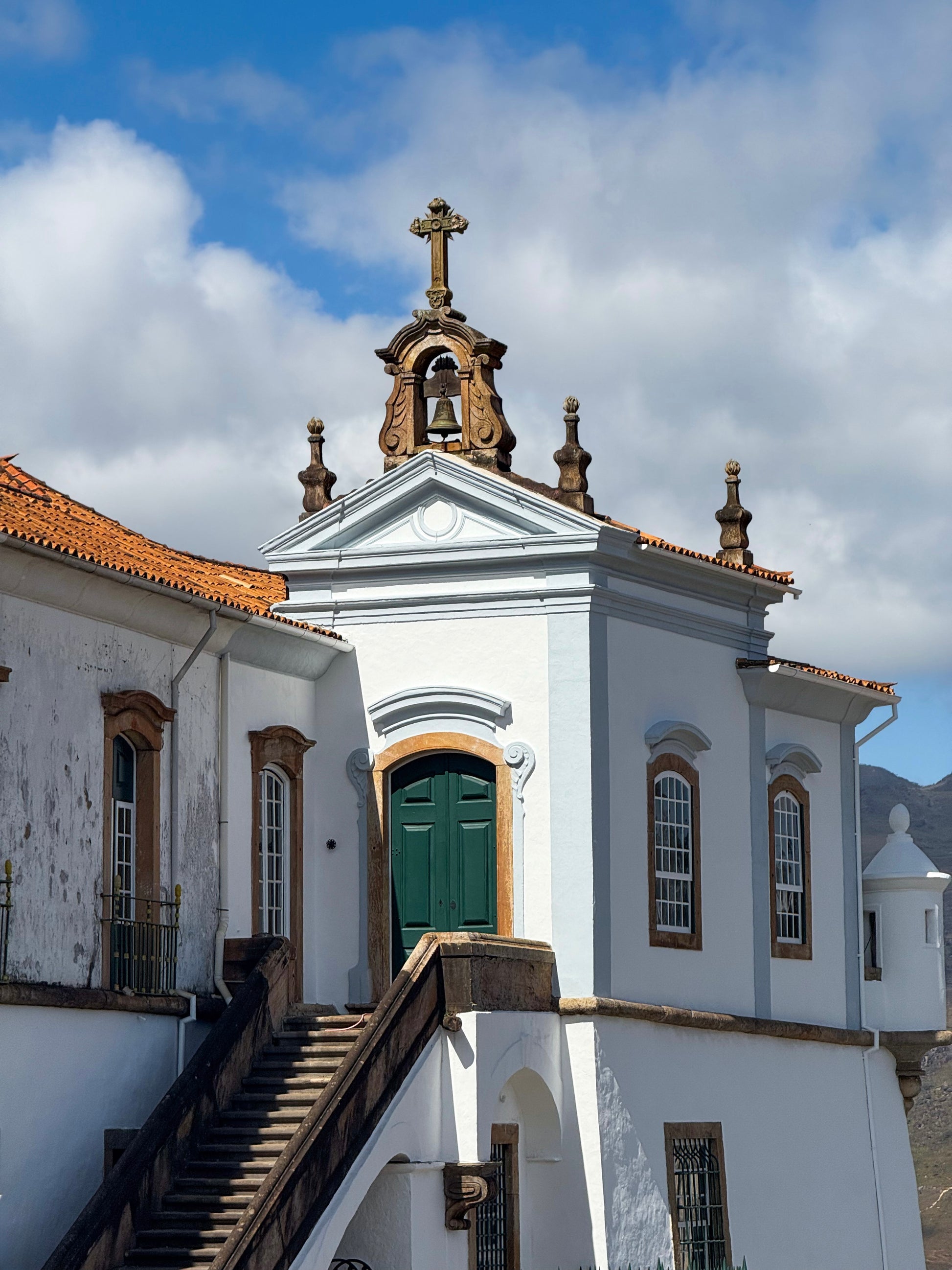 The Historic Baroque Church in Ouro Preto by Leslie Brashear Photography features green doors, windows, a cross-topped white exterior, and an outdoor staircase—capturing the city's colonial charm beneath a vibrant blue sky.
