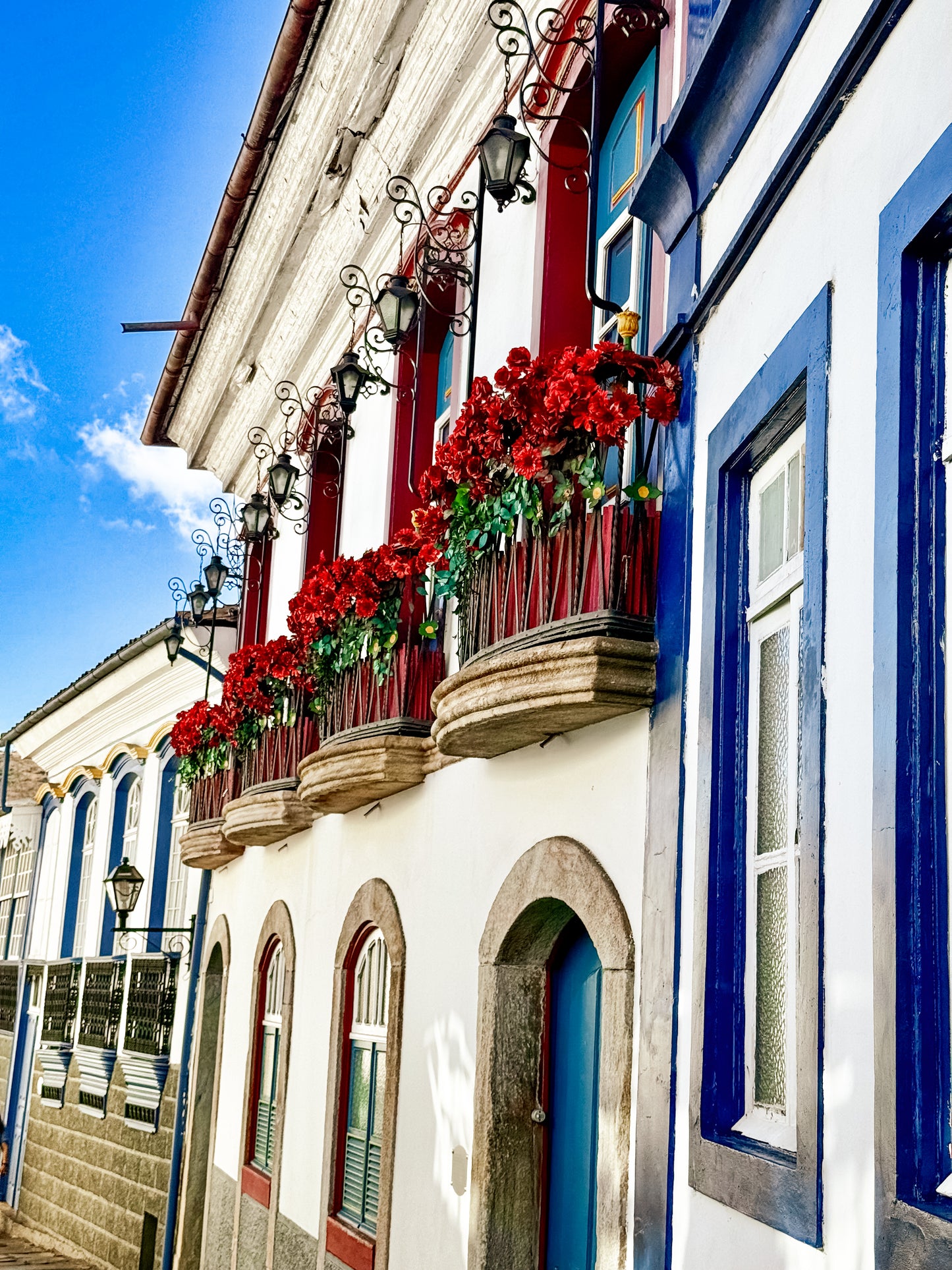 Charming Balcony in Ouro Preto with Flower Boxes by Leslie Brashear Photography captures a vibrant facade with red flowers, wrought iron balconies, white walls, and blue-trimmed windows under a clear sky.