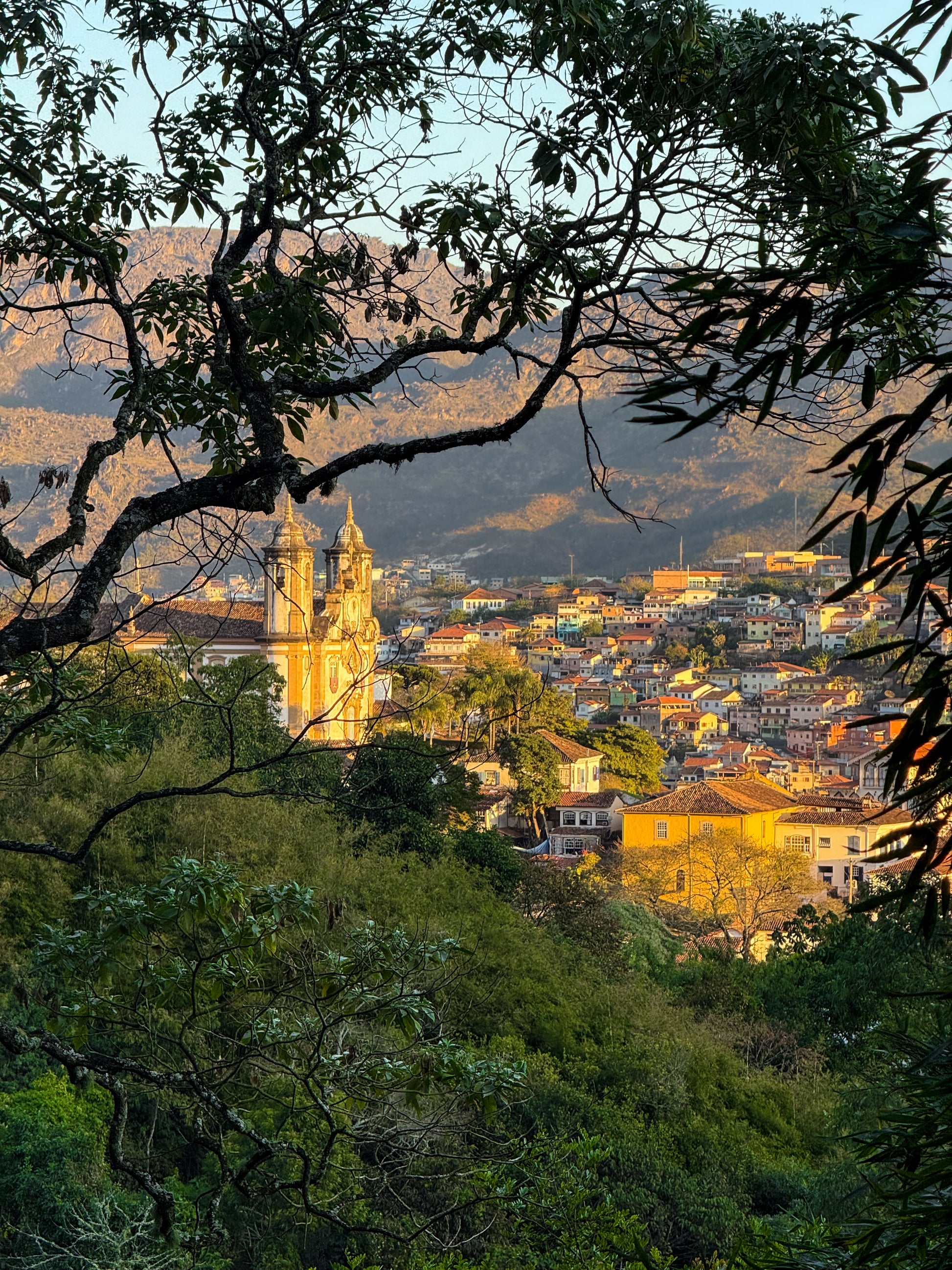 Charming Ouro Preto at Dusk by Leslie Brashear Photography captures the colonial architecture, colorful hillside buildings, and a church framed by tree branches, with mountains in the background.