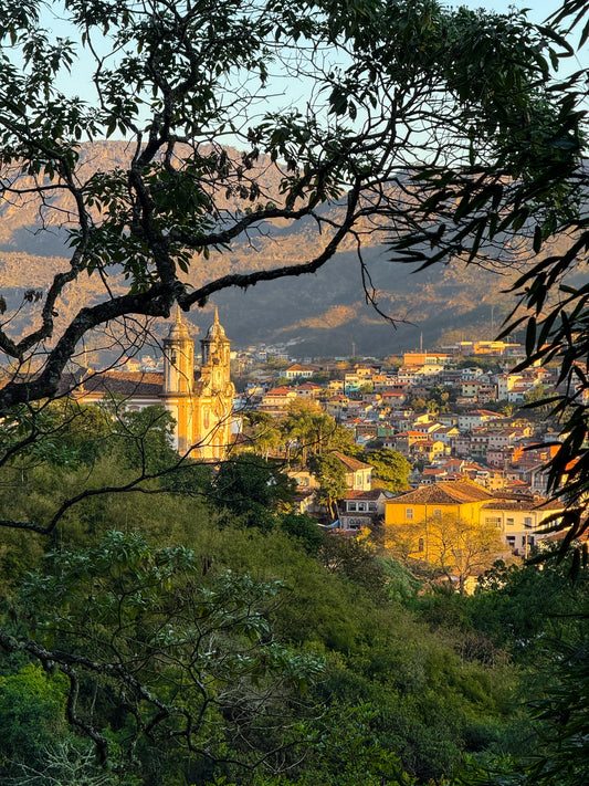 Charming Ouro Preto at Dusk by Leslie Brashear Photography captures the colonial architecture, colorful hillside buildings, and a church framed by tree branches, with mountains in the background.