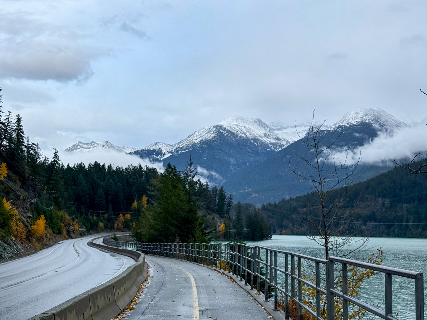 The "Snowy Mountain Highway - Fine Art Print" by Leslie Brashear Photography captures a winding road and bike path beside a serene lake, set against snow-capped mountains, forested hills, and dramatic British Columbia skies.