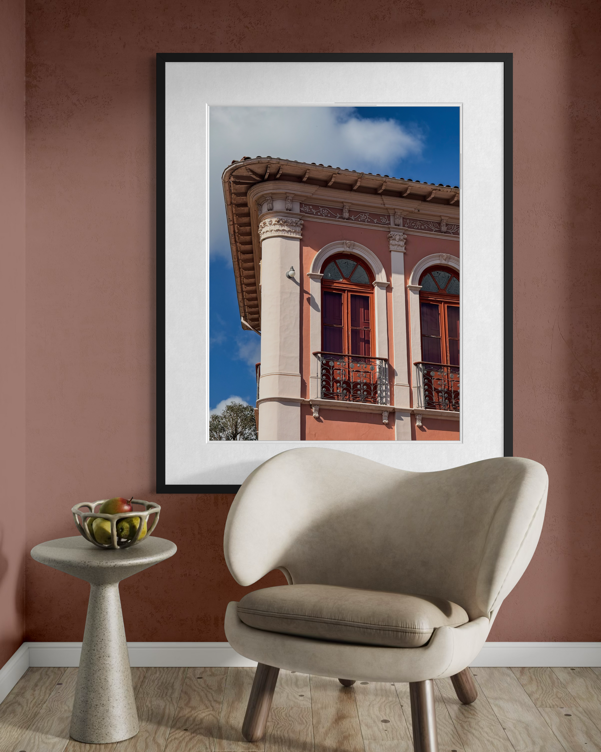 A beige chair and small table with fruit sit in front of Leslie Brashear Photography’s “Ornate Balcony with Arched Windows,” featuring Ouro Preto’s historic architecture.