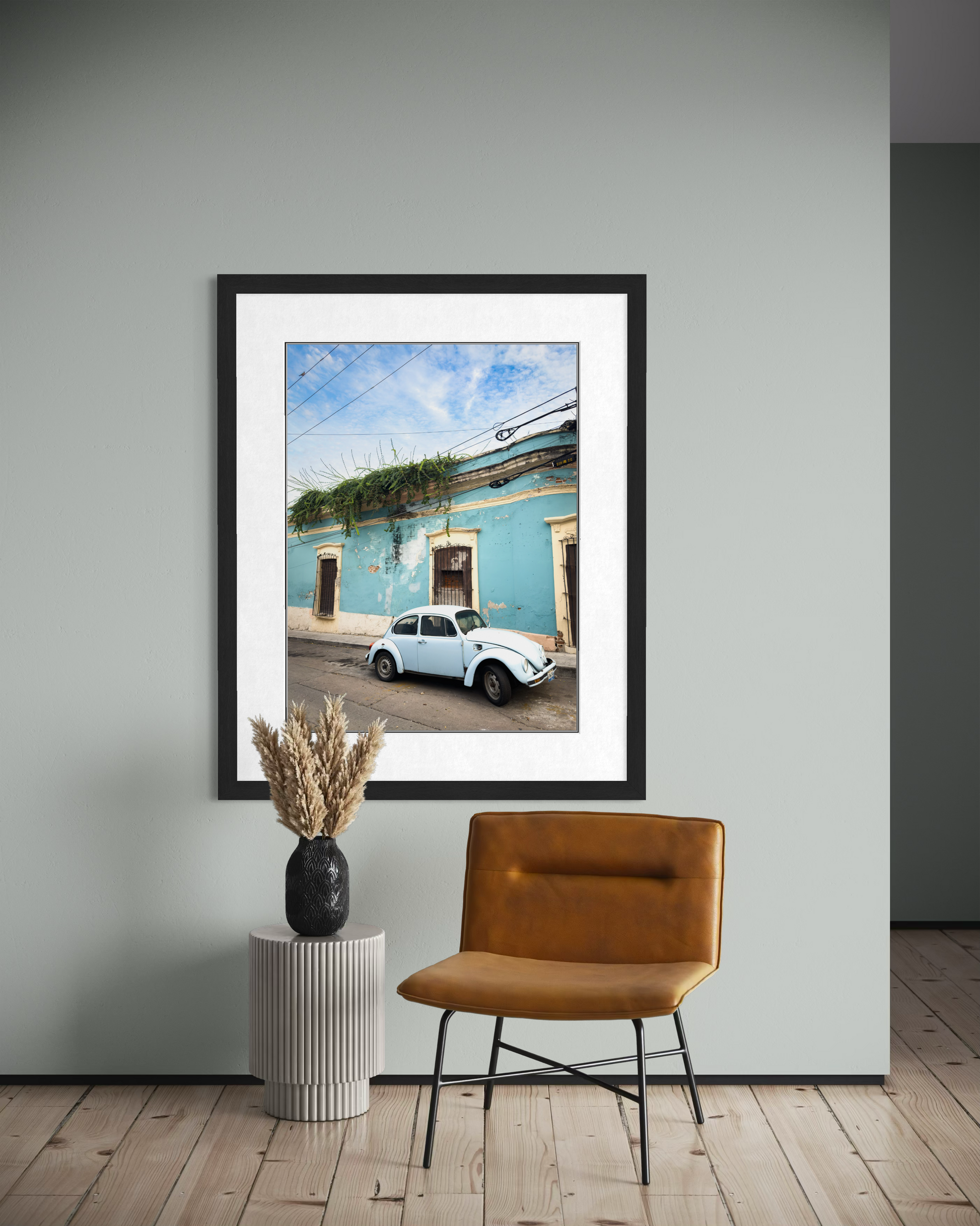 The "Vintage VW Beetle Parked on a Side Street" print by Leslie Brashear Photography hangs above a small table with a vase of dried plants and a tan chair, displayed on a pale wall.
