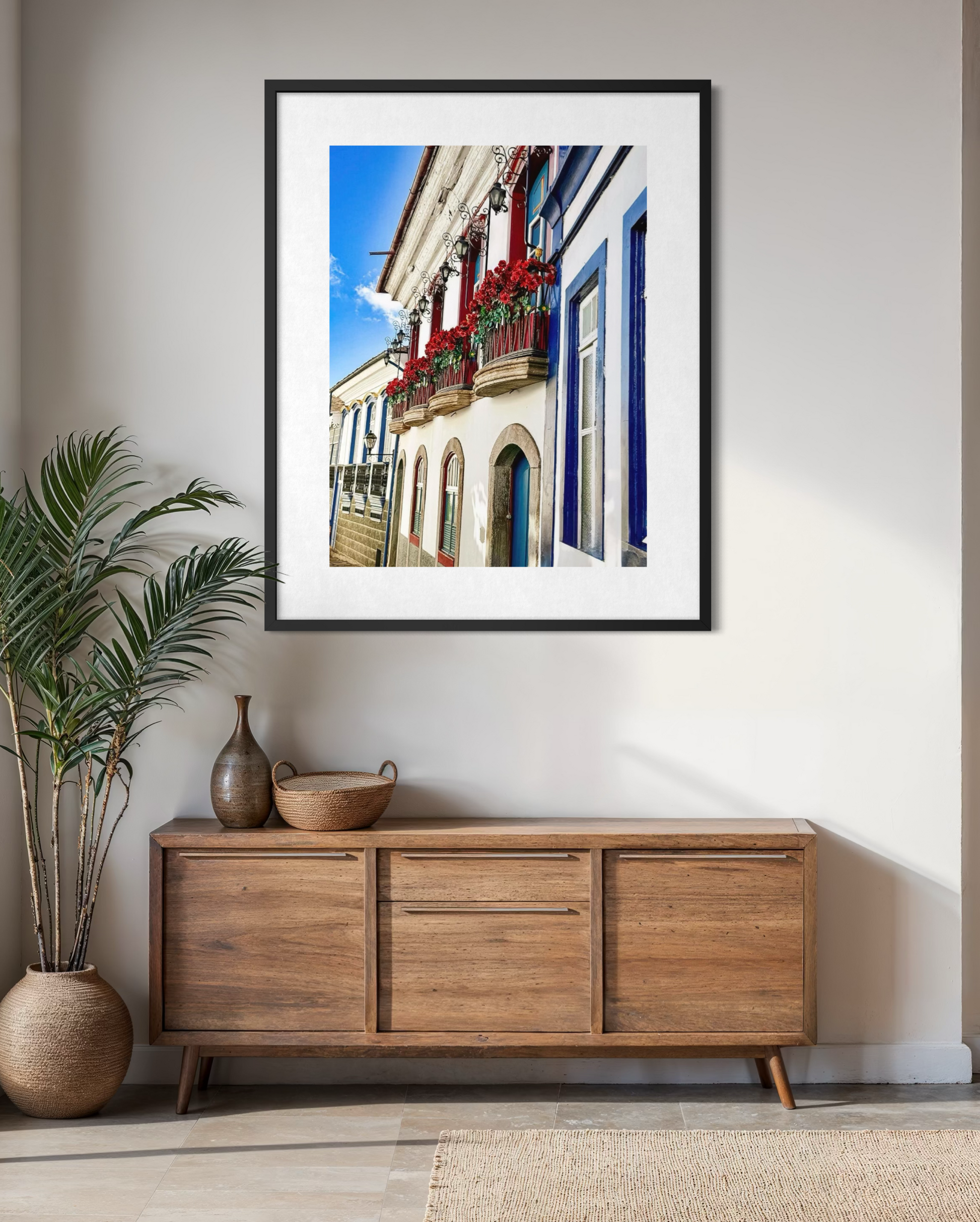 “Charming Balcony in Ouro Preto with Flower Boxes” by Leslie Brashear Photography, featuring vibrant colonial architecture and red flowers, is framed and displayed above a wooden sideboard in a modern, minimalist room.