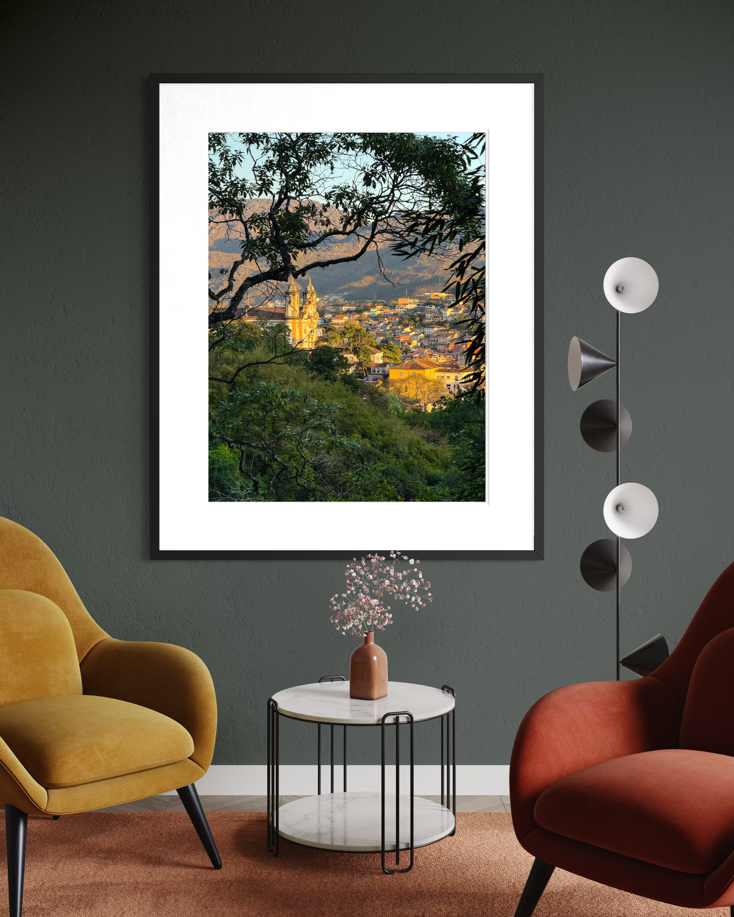 “Charming Ouro Preto at Dusk” by Leslie Brashear Photography, a framed print of sunlit churches and colonial architecture, hangs on a dark wall above a round white table between mustard yellow and rust orange armchairs.