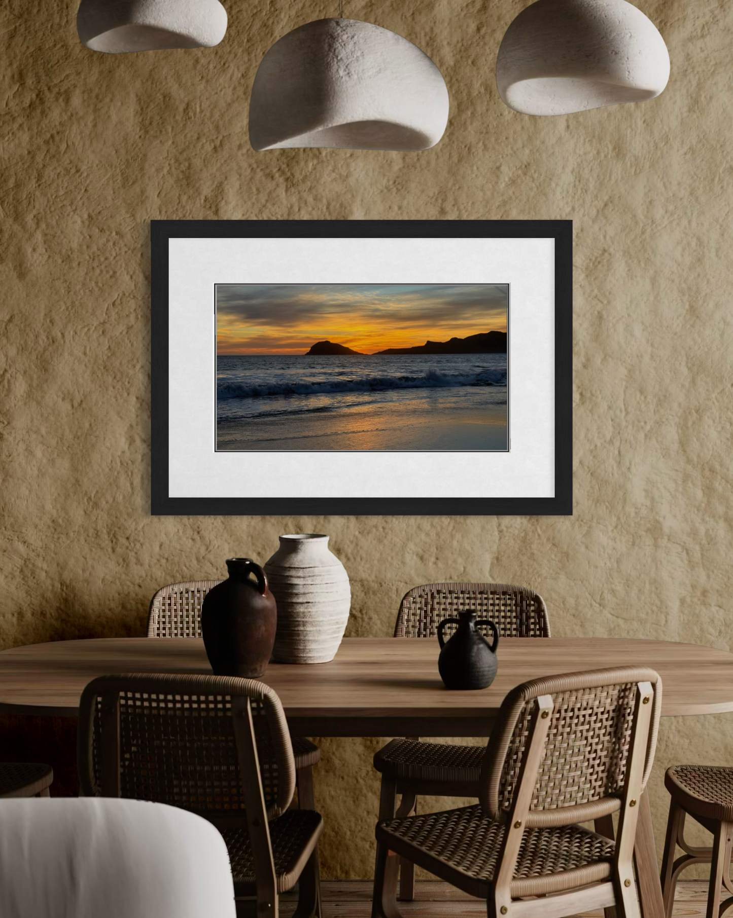 “Sunset Over the Coast of Mazatlan” by Leslie Brashear Photography, a framed photo of a vivid sky and ocean at sunset, hangs on a beige wall above a wooden dining table with woven chairs and ceramic vases.