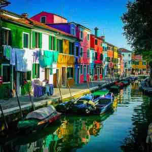 Colorful Burano - Fine Art Print features a vibrant canal scene with colorful buildings, small boats, and laundry drying in the bright daylight.