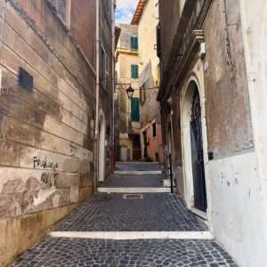 Passage Through Tivoli shows a narrow cobblestone alleyway leading through an elegant stone gateway between tall, weathered buildings in an old European town.